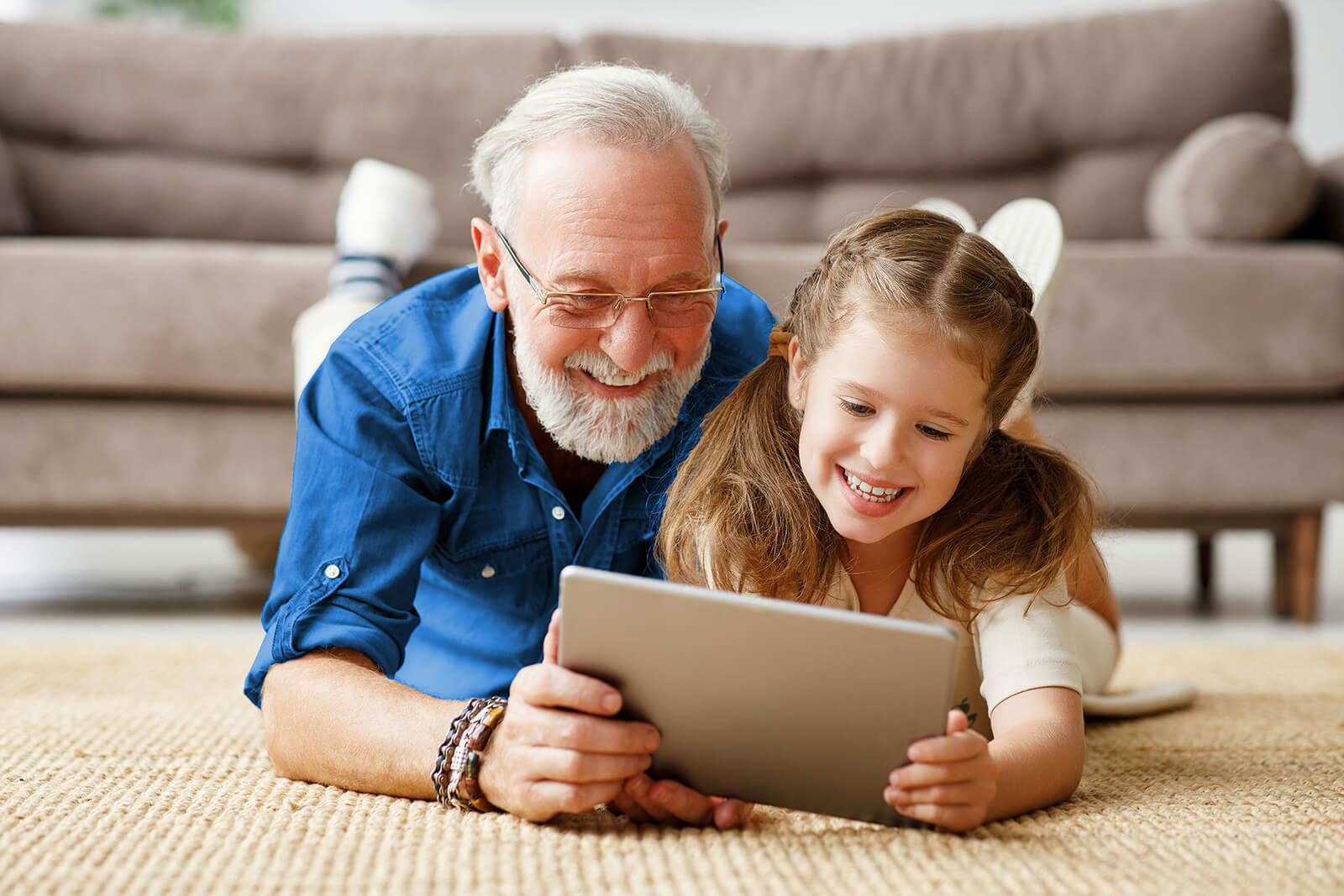 A smiling grandfather and granddaughter lie belly-down on the carpet in front of a sofa, each holding one side of a tablet.