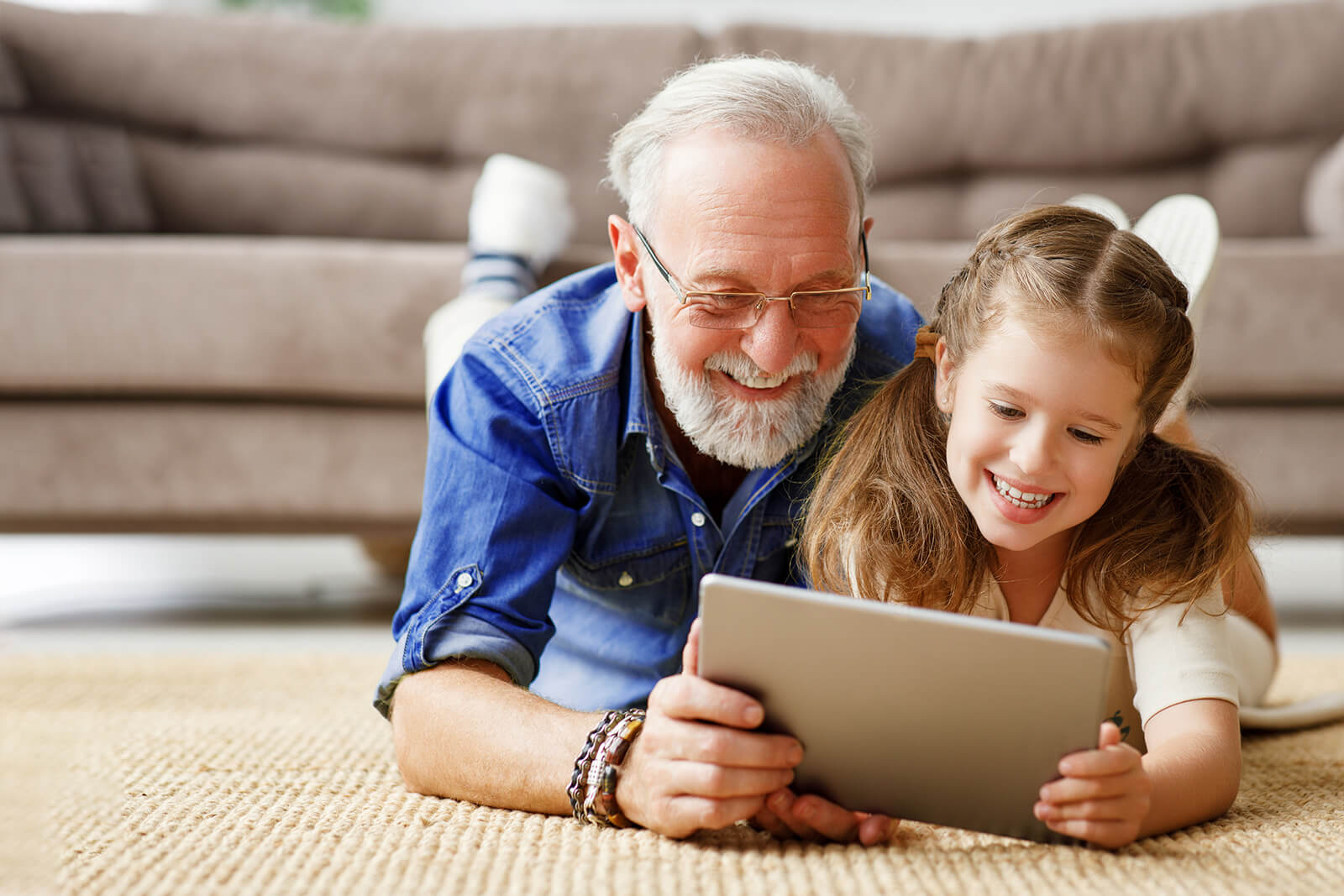 Un grand-père et sa petite-fille sont couchés, tout sourire et à plat ventre, sur un tapis en face d’un sofa, chacun tenant par le côté la même tablette.