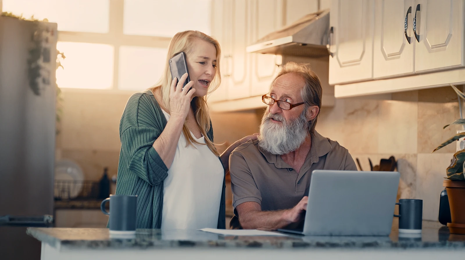 Two people in a kitchen, with one seated at a laptop and the other standing nearby while talking on a phone.