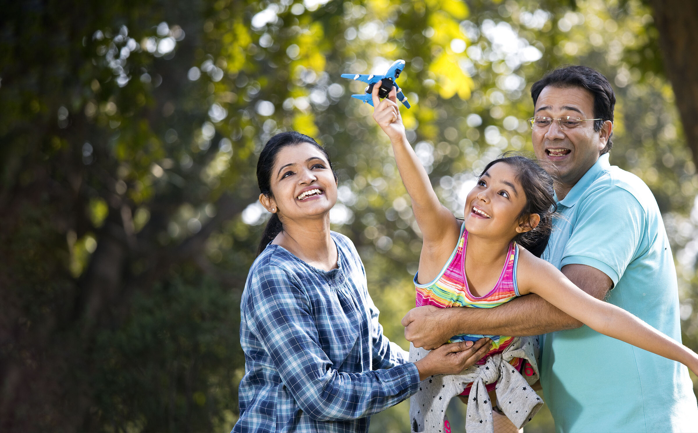 A smiling couple hold their daughter in their arms while she lifts a toy plane up to the sky, surrounded by trees.