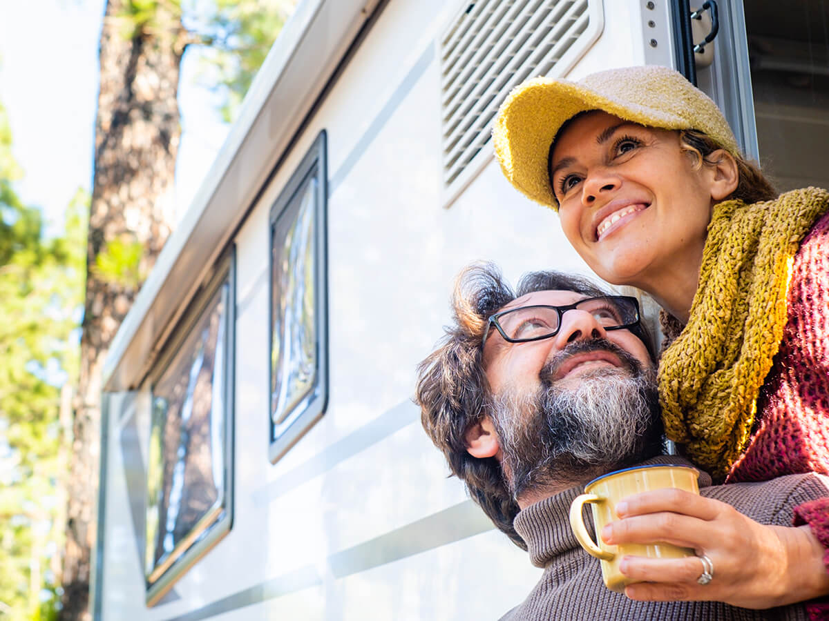 In a sunny, wooded area, a smiling couple stand in the door of a motorhome. The woman holds a coffee mug.