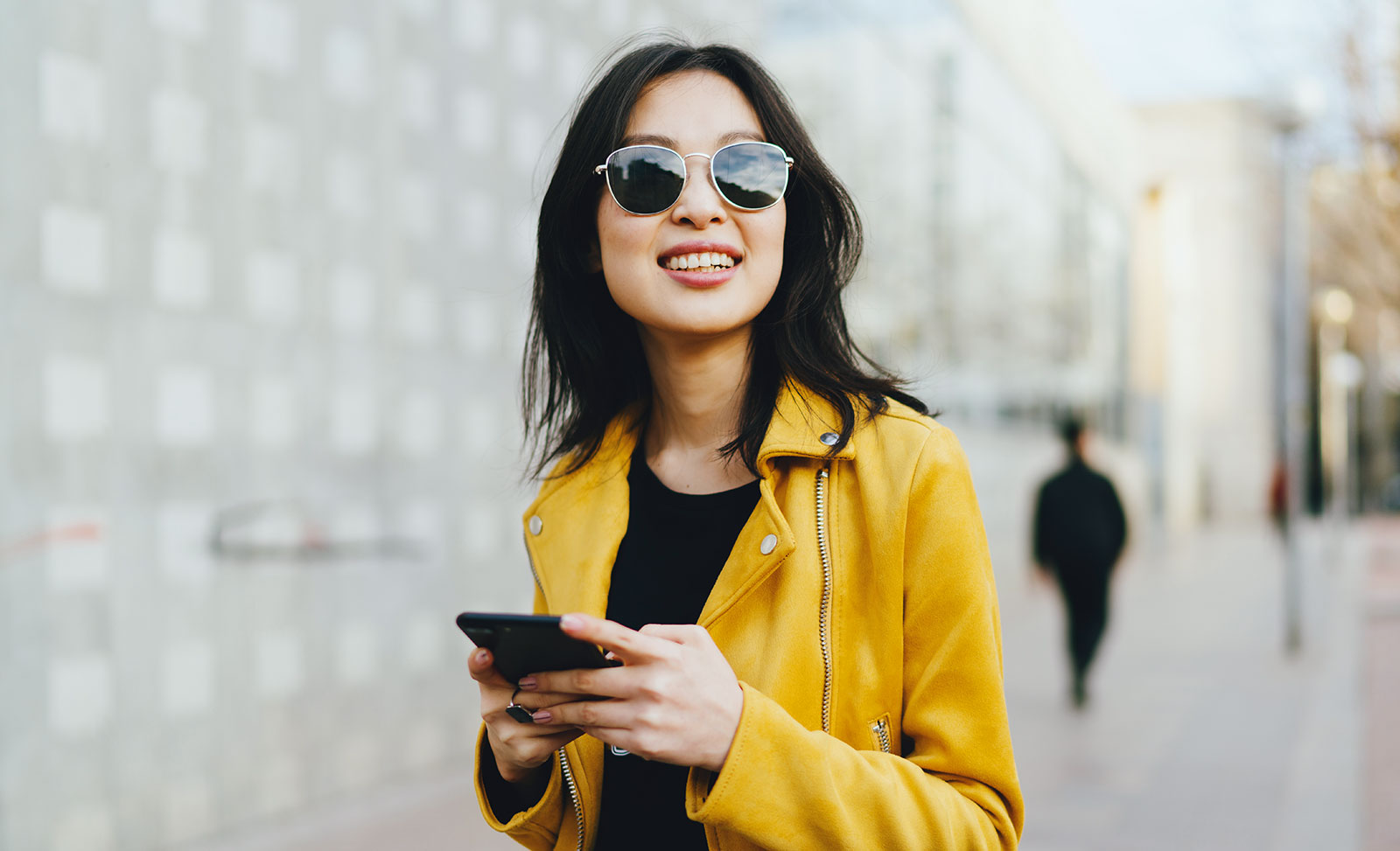 A young woman in sunglasses and yellow leather jacket texting on her phone.