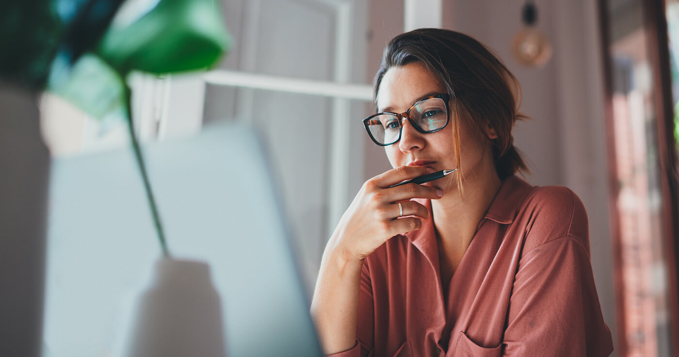 Une femme portant des lunettes est assise chez elle. Elle tient un stylo à la main avec un air pensif.