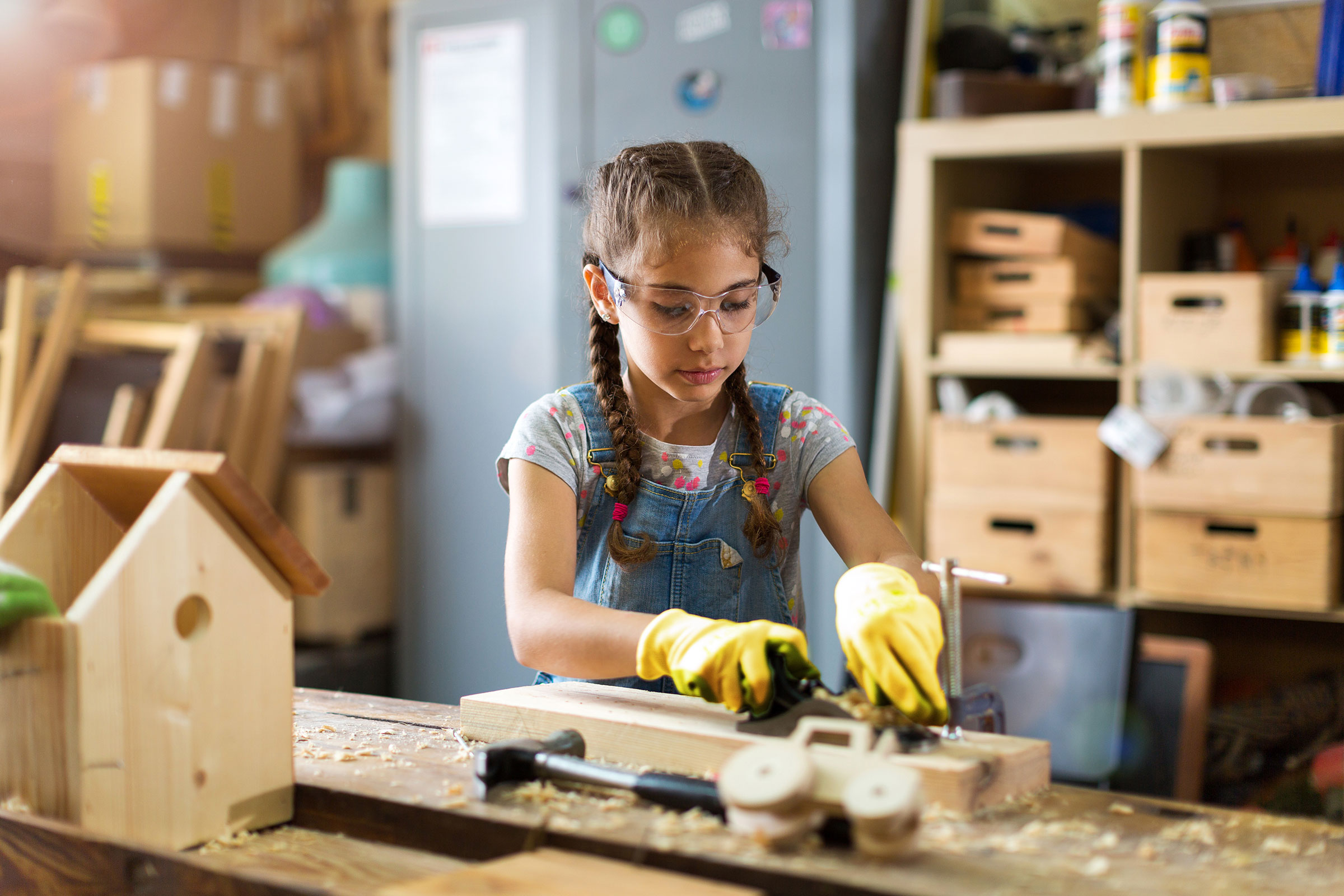 A young girl wearing safety glasses and gloves carves wooden toys on a work bench.