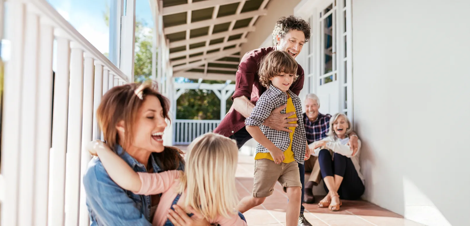 A multi-generational family gathered on a cozy porch. Grandparents sit together as a couple plays with their young children.