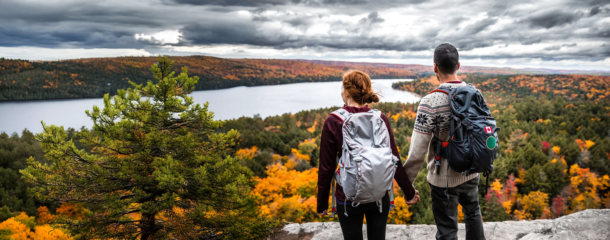 Couple de randonneurs tenant la main devant un paysage d’automne avec un lac et des forêts colorées.