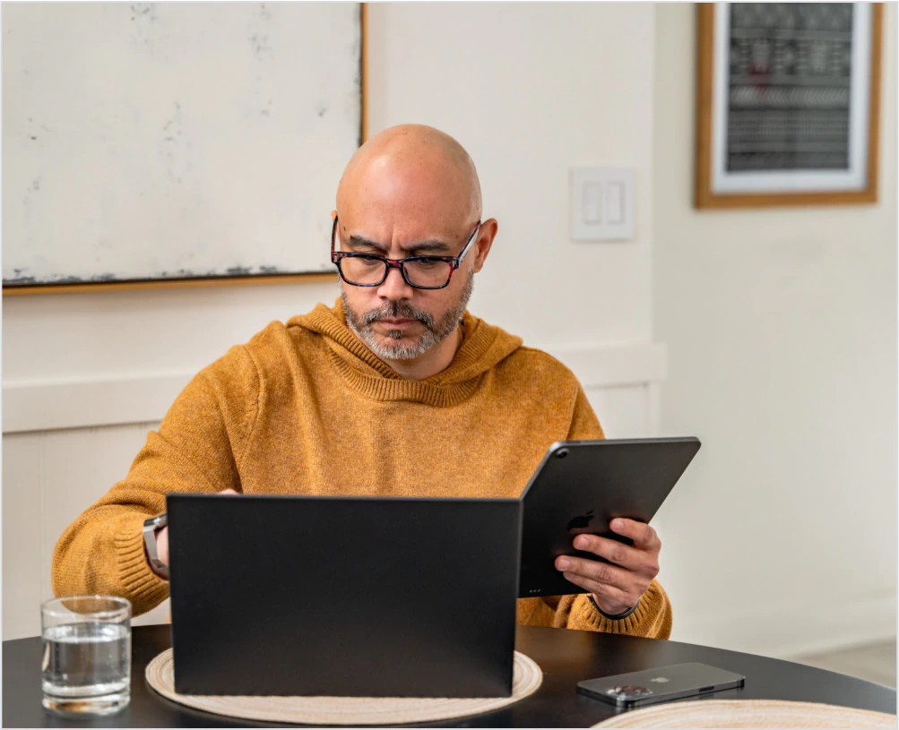 Homem sentado à mesa usando um tablet e um laptop para gerenciar o acesso com o 1Password XAM
