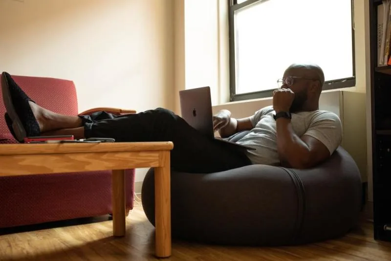 A man reclines in a soft chair with his legs up on a coffee table looking at a laptop