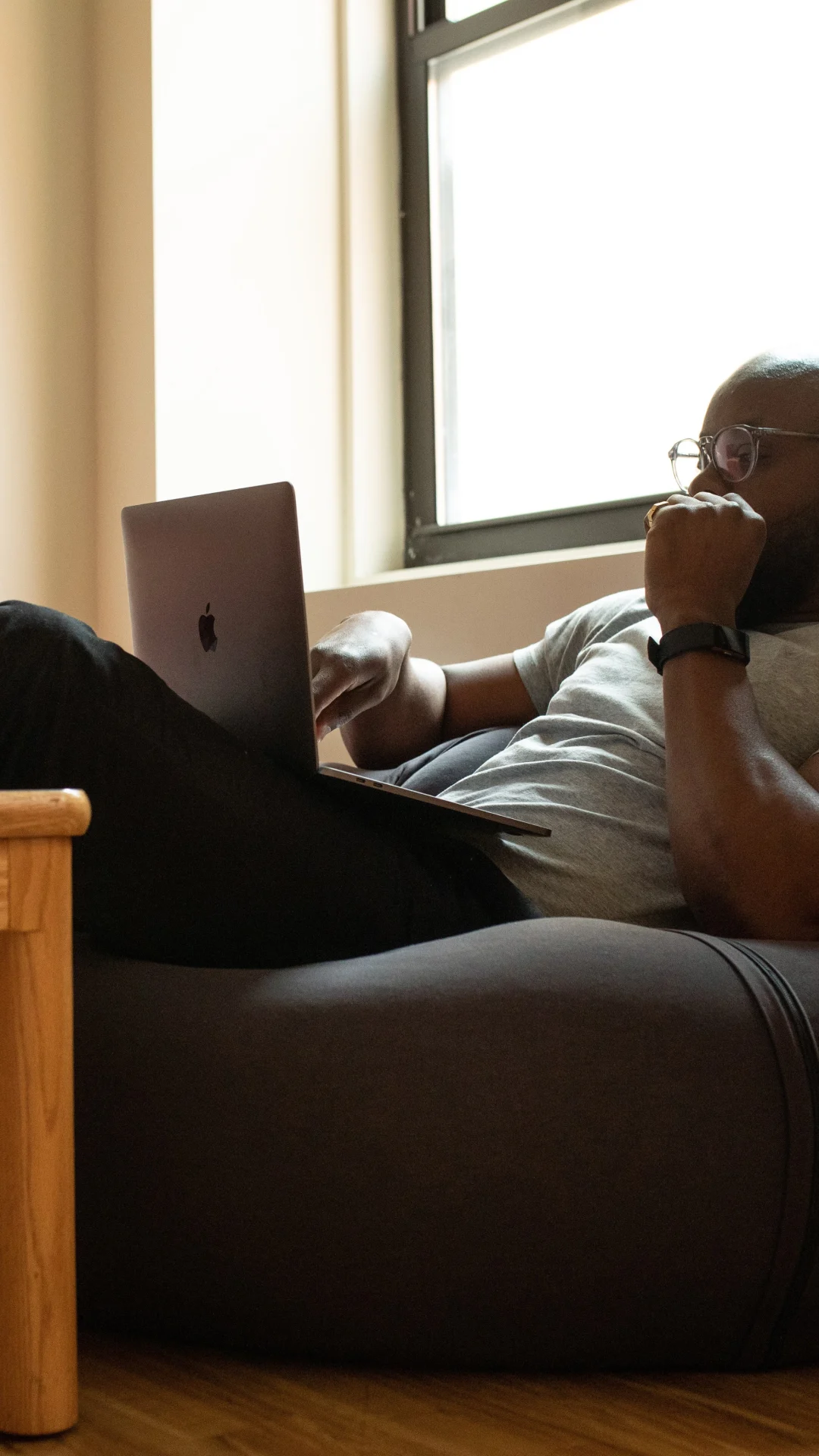 A man in white t-shirt sitting on a floor pillow with a laptop in his lap.