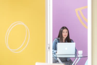 Woman sitting behind a laptop Woman sitting in front of yellow and pink walls with laptop