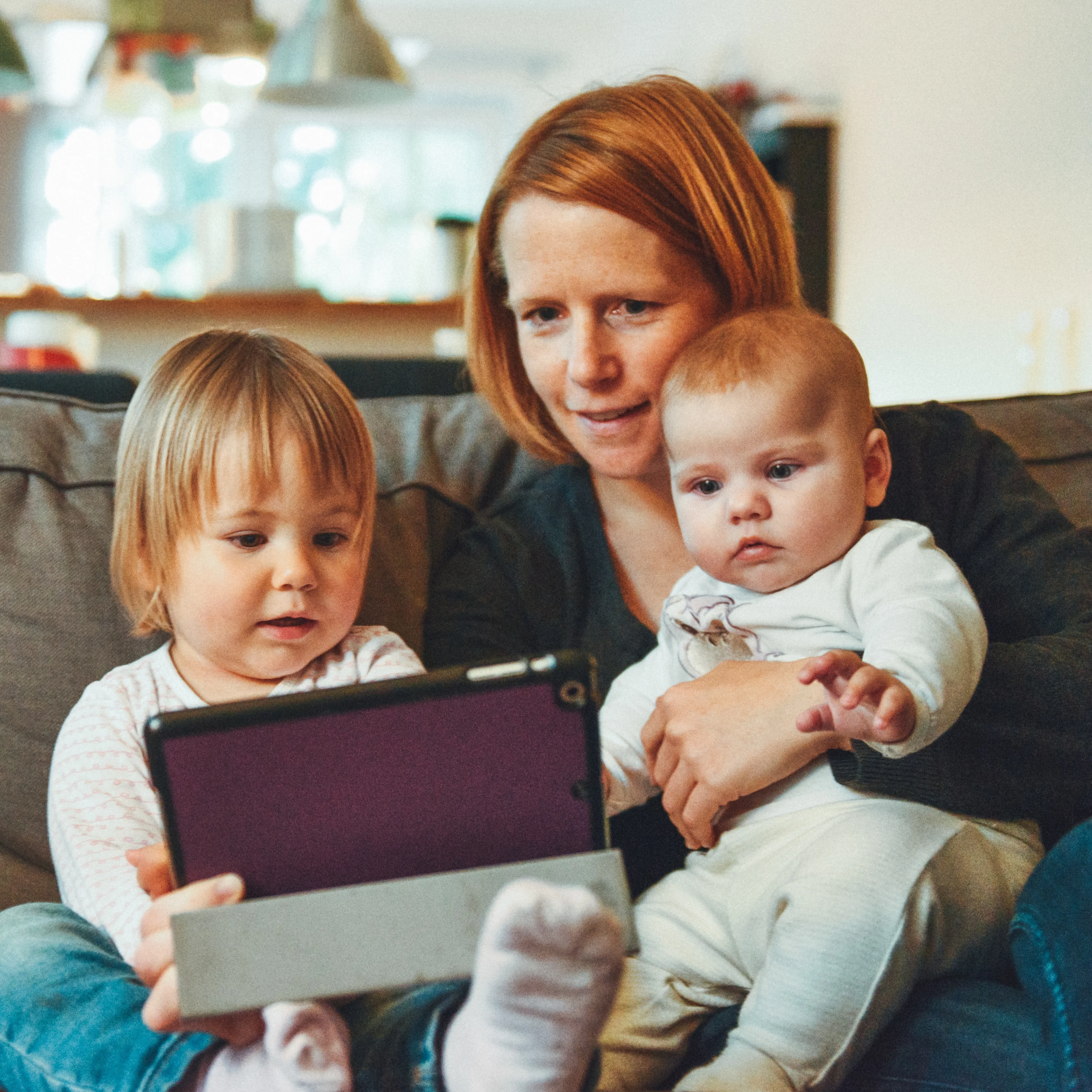 Woman sitting in couch with small child and baby looking at a tablet together