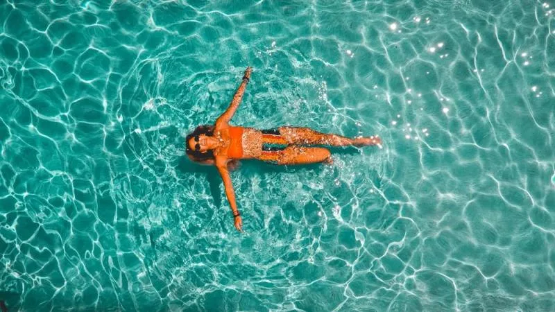A woman in a bathing suit lies in a clear blue swimming pool looking up at the camera