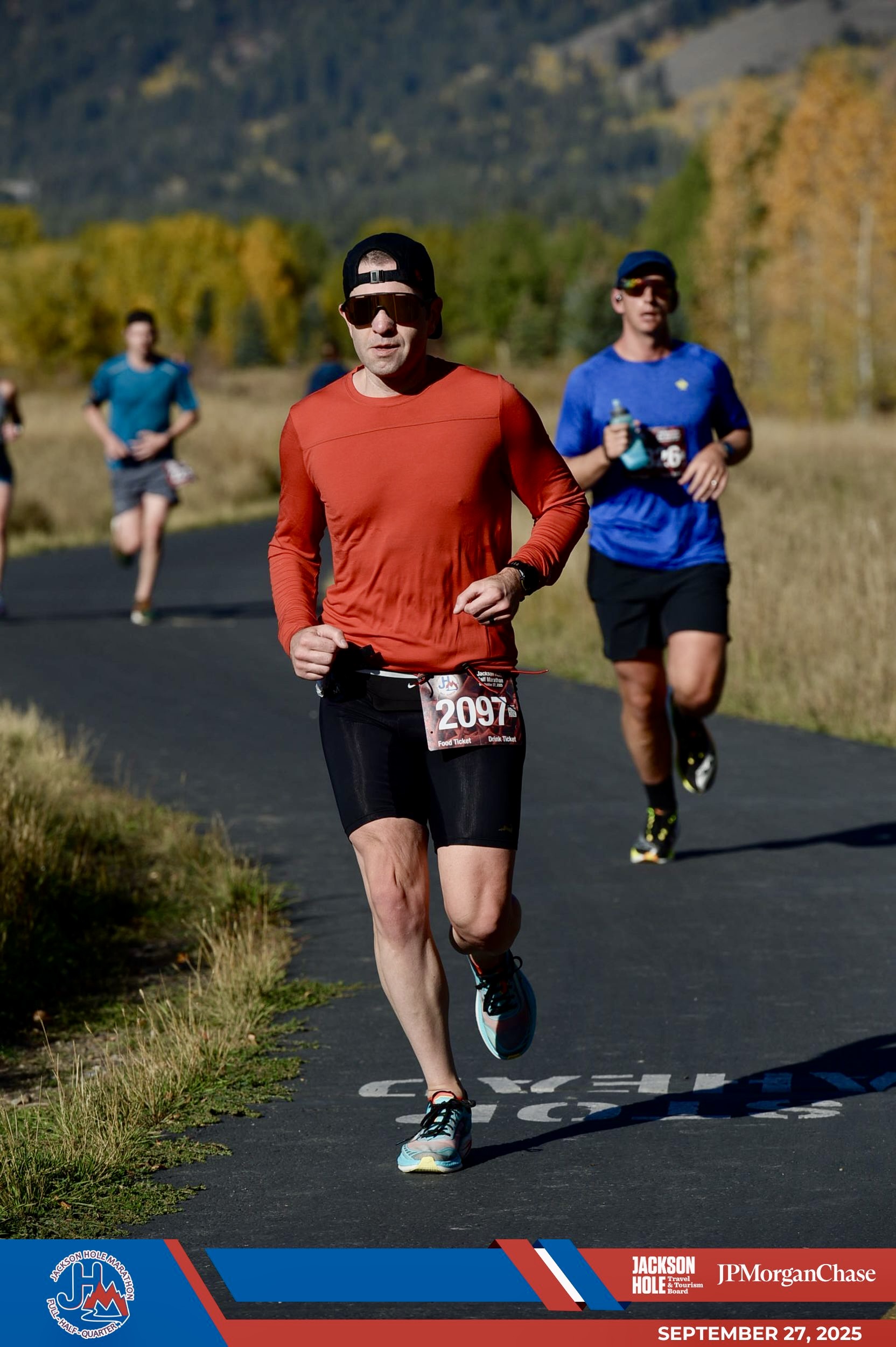Guillermo Esteves, correndo por um caminho curvo pavimentado ladeado por gramíneas douradas e árvores com folhagens de outono ao fundo, durante a Meia Maratona Hole. Ele está usando um chapéu preto, óculos escuros dourados, camisa vermelha de manga comprida, meia-calça preta, sapatos azuis claros e um colete de corrida número 2097 em um cinto de corrida. Três outros corredores podem ser vistos atrás dele. Os gráficos em azul e vermelho na parte inferior exibem os logotipos da Jackson Hole Marathon Races, Jackson Hole Travel & Tourism Board e JP Morgan Chase, junto com a data, 27 de setembro de 2025.