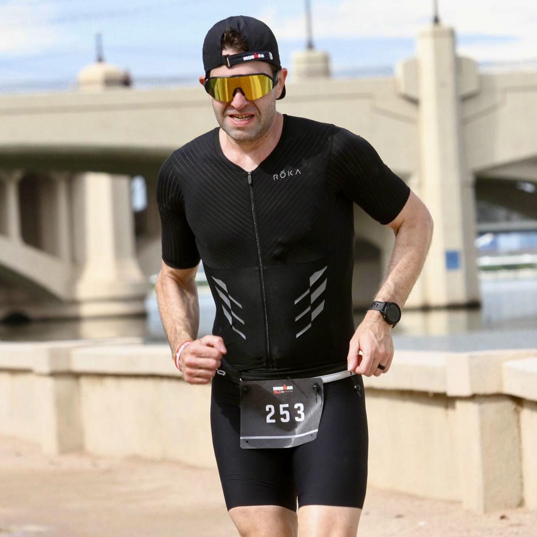 Me, running on a dirt path, with the Mill Avenue Bridge in Tempe in the background. I'm wearing a black running hat backwards, gold sunglasses, black tri suit, and orange shoes.