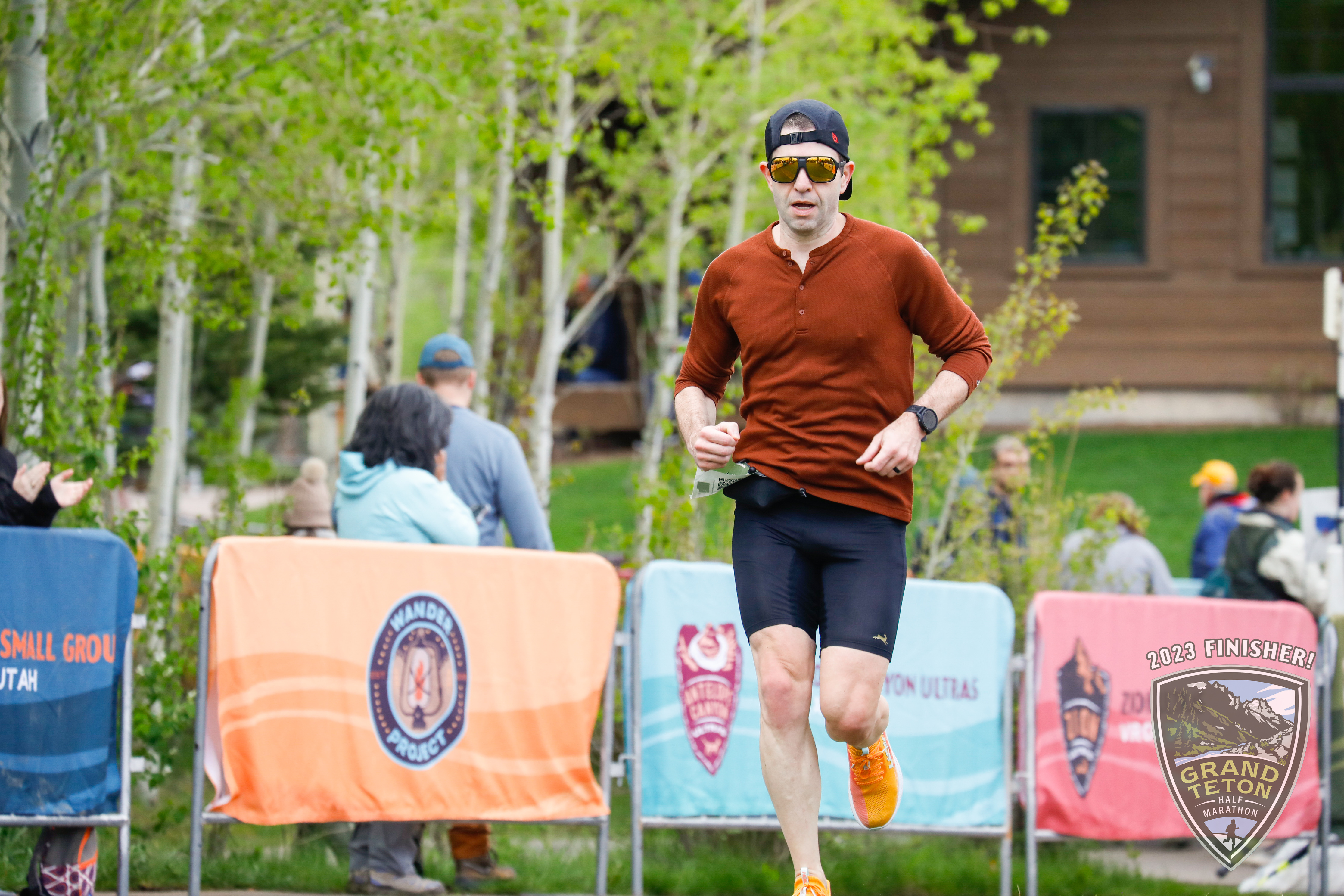 Me, mid-stride at the finish chute of the Grand Teton Half Marathon. I'm wearing a black running hat, gold sunglasses, a red henley, black tights, and orange shoes.