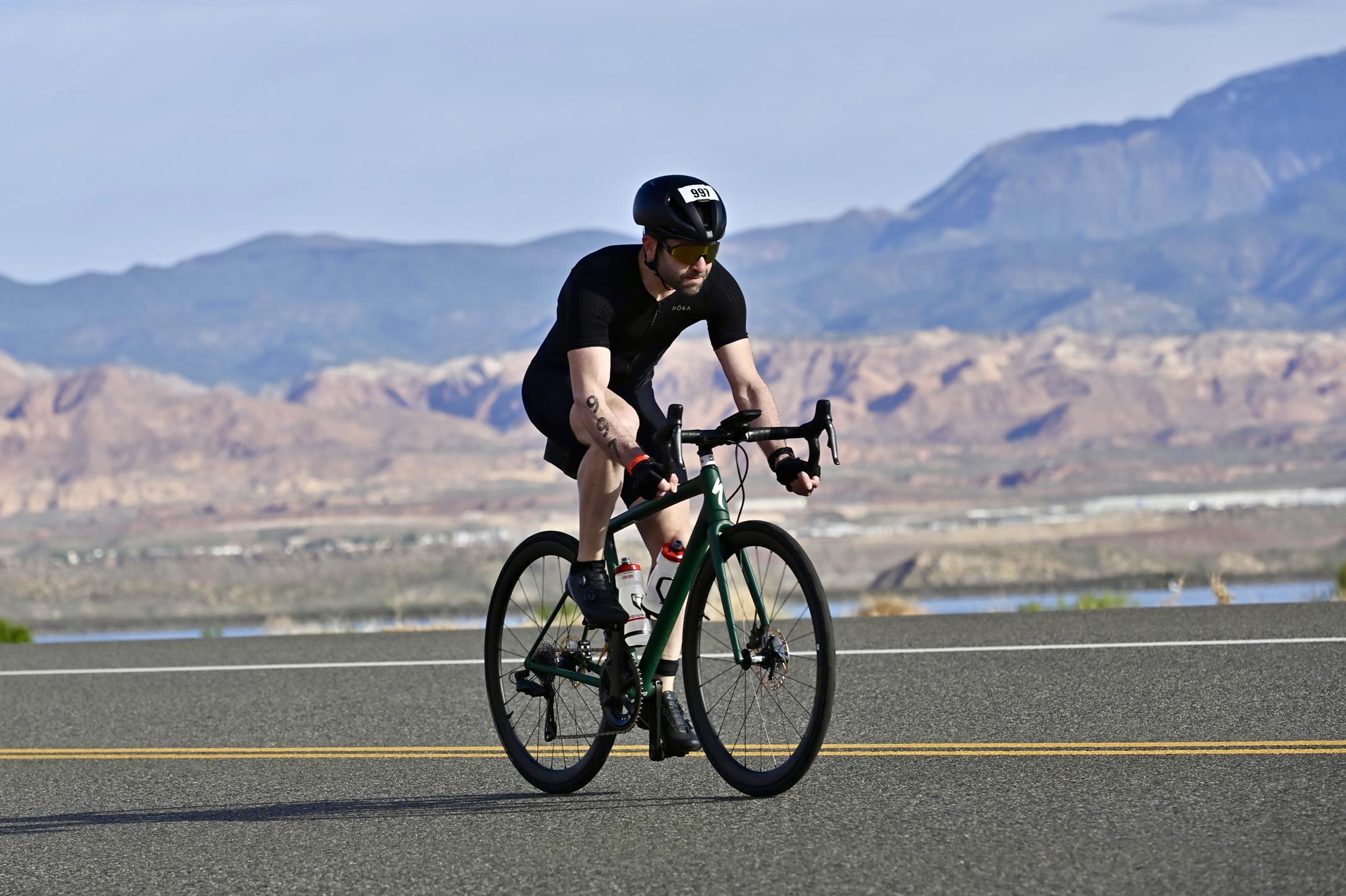 Me, riding a green Specialized Aethos bike through SR7 in southern Utah. Mountains can be seen in the background, along with a little bit of the Sand Hollow reservoir. I'm wearing a black helmet, black trisuit, black gloves, black shoes, and gold sunglasses. I have a race tattoo in my right arm with the number 997.