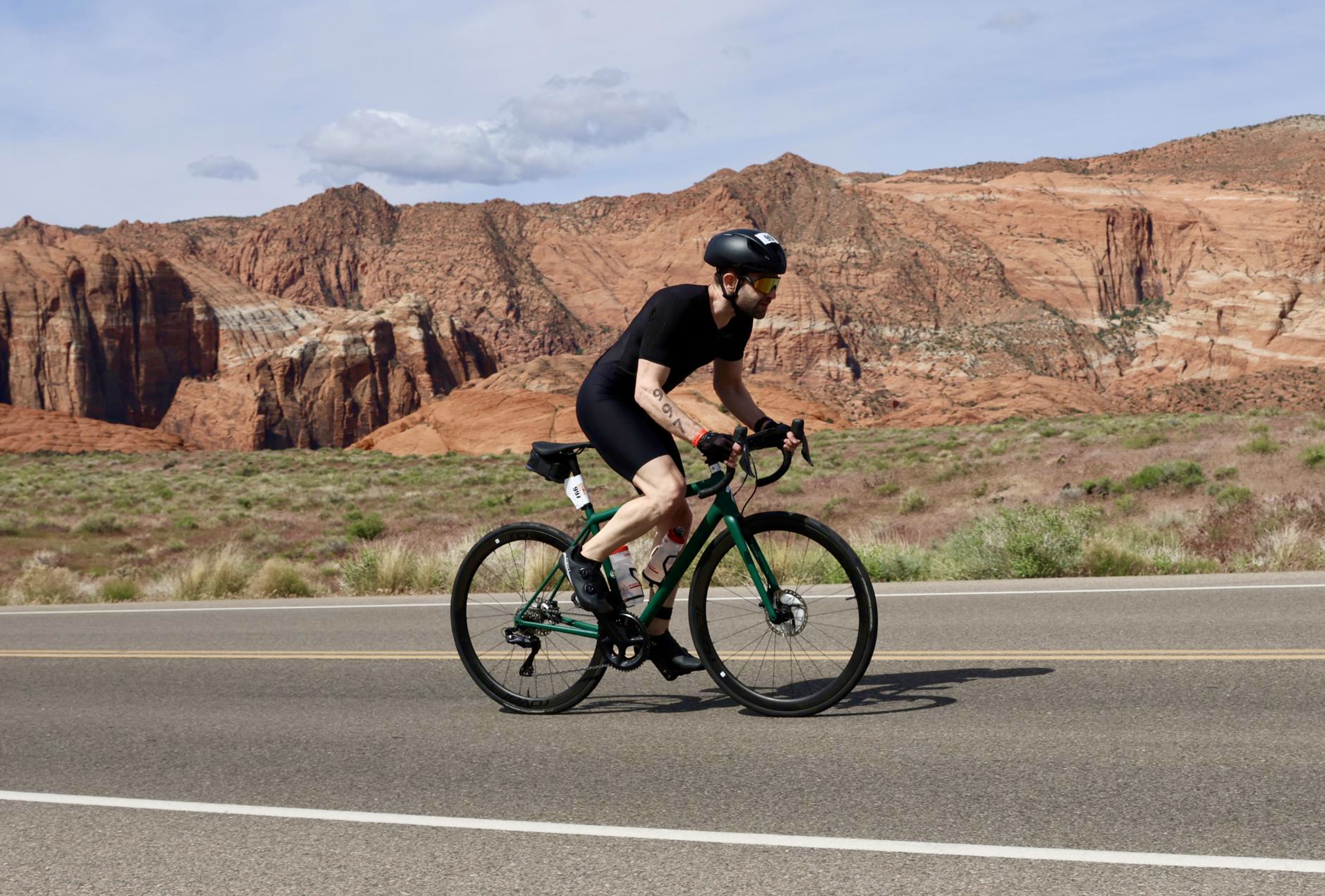 Me, climbing off the saddle on a green Specialized Aethos bike, through Snow Canyon, with its red cliffs visible in the background. I'm wearing a black helmet, black trisuit, black gloves, black shoes, and gold sunglasses. I have a race tattoo in my right arm with the number 997.