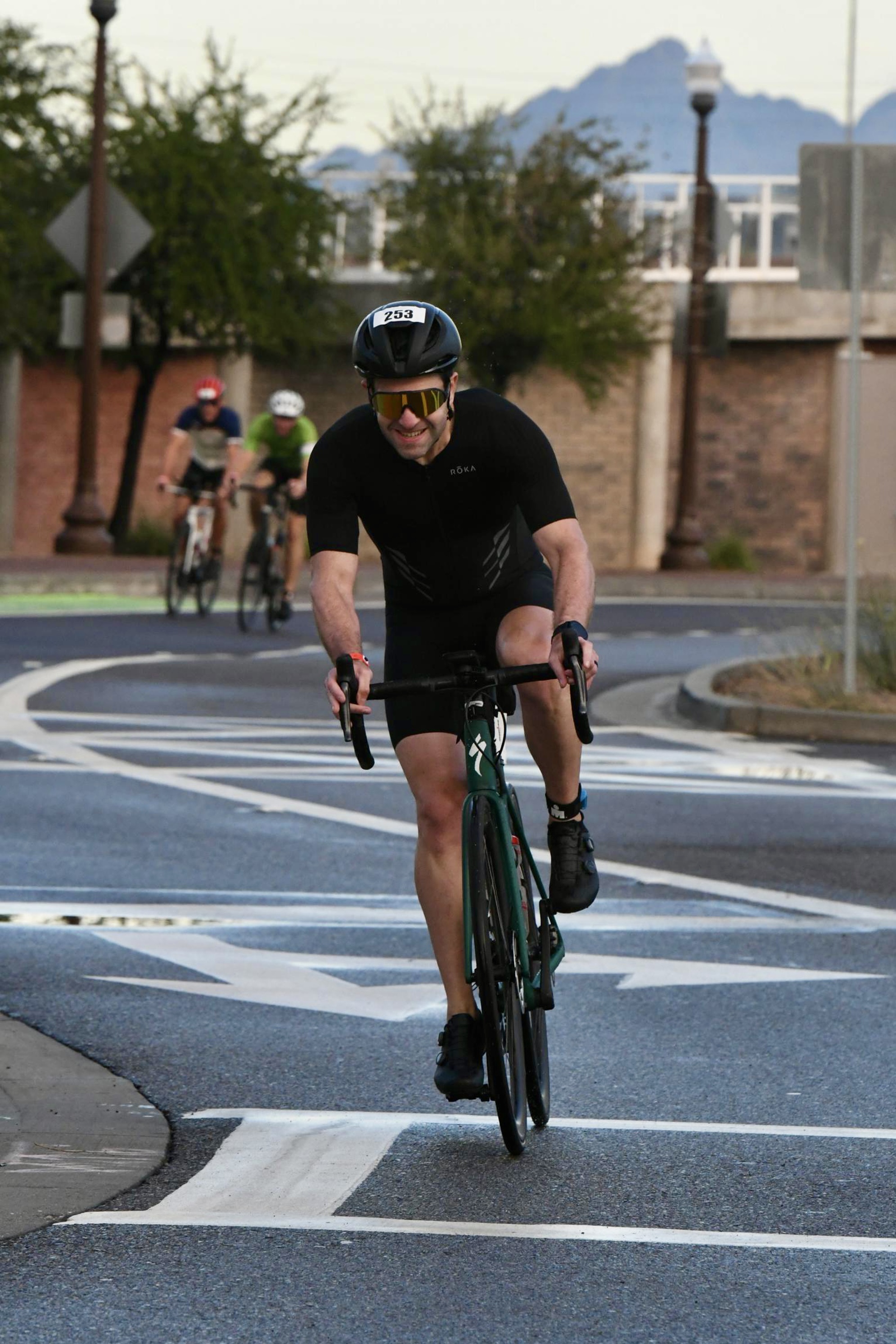 Me, riding a green Specialized Aethos bike around a roundabout, with wet pavement. I'm wearing a black helmet, gold sunglasses, black tri suit, and black shoes. Behind me, two other riders.