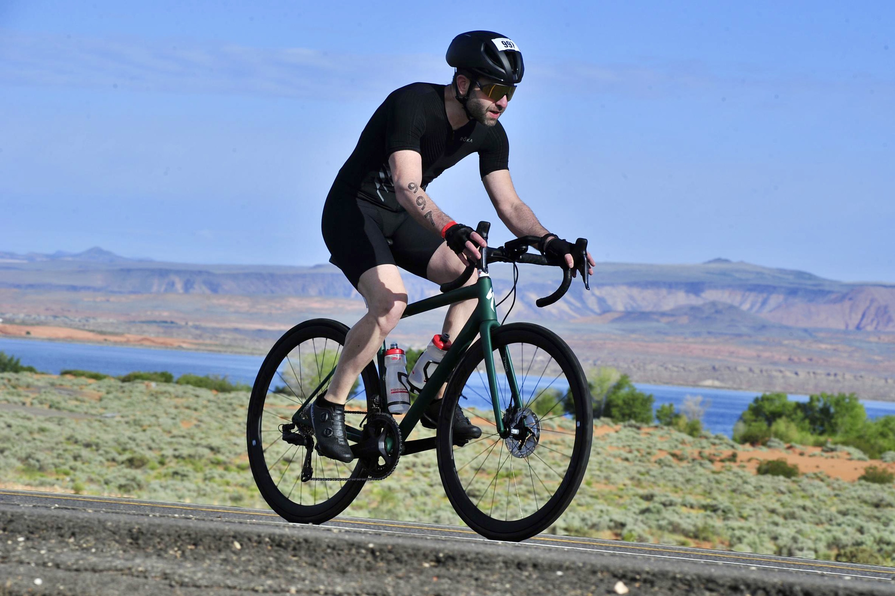 Me, riding a green Specialized Aethos bike through SR7 in Utah. The Sand Hollow reservoir and mountains can be seen in the background, under clear blue skies. I'm wearing a black helmet, black trisuit, black gloves, black shoes, and gold sunglasses. I have a race tattoo in my right arm with the number 997.