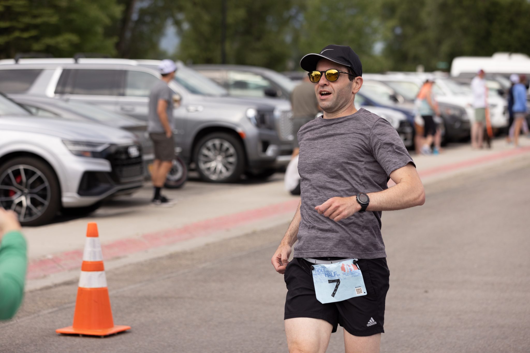 Guillermo Esteves, seen mid-stride while arriving at the finish line of the Jackson Hole Marathon, and wearing a black cap, gold sunglasses, gray t-shirt, black shorts, and a race bib with the number 7.