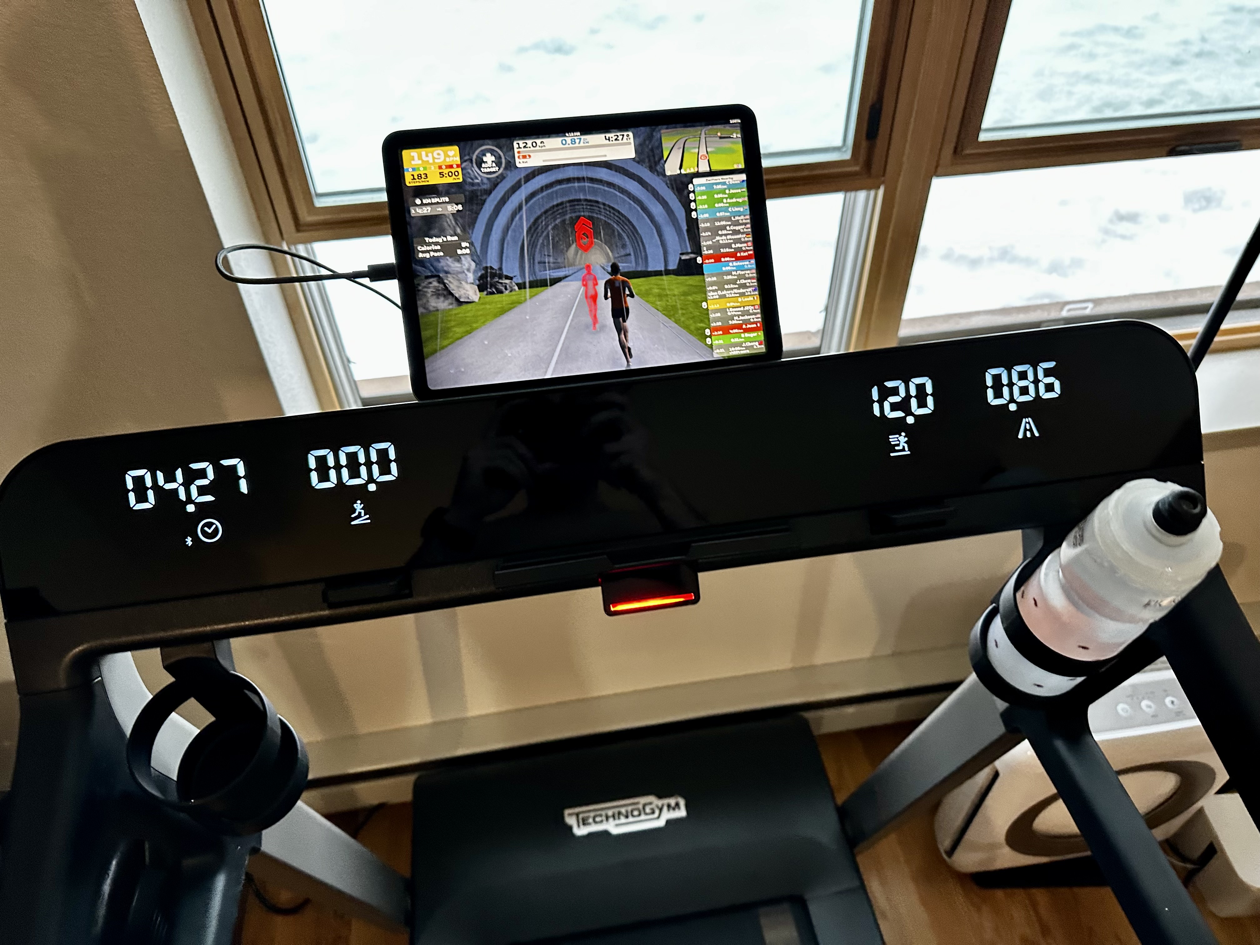 The console of a Technogym MyRun treadmill, from the point of view of someone running on it. The display shows 4:27 minutes elapsed, 0% incline, 12.0 km/h, and 0.86 km. On top of the console, a connected iPad is running Zwift. A water bottle is on the right bottle holder. The treadmill is in front of the windows; a heavy cover of snow can be seen on the ground outside.