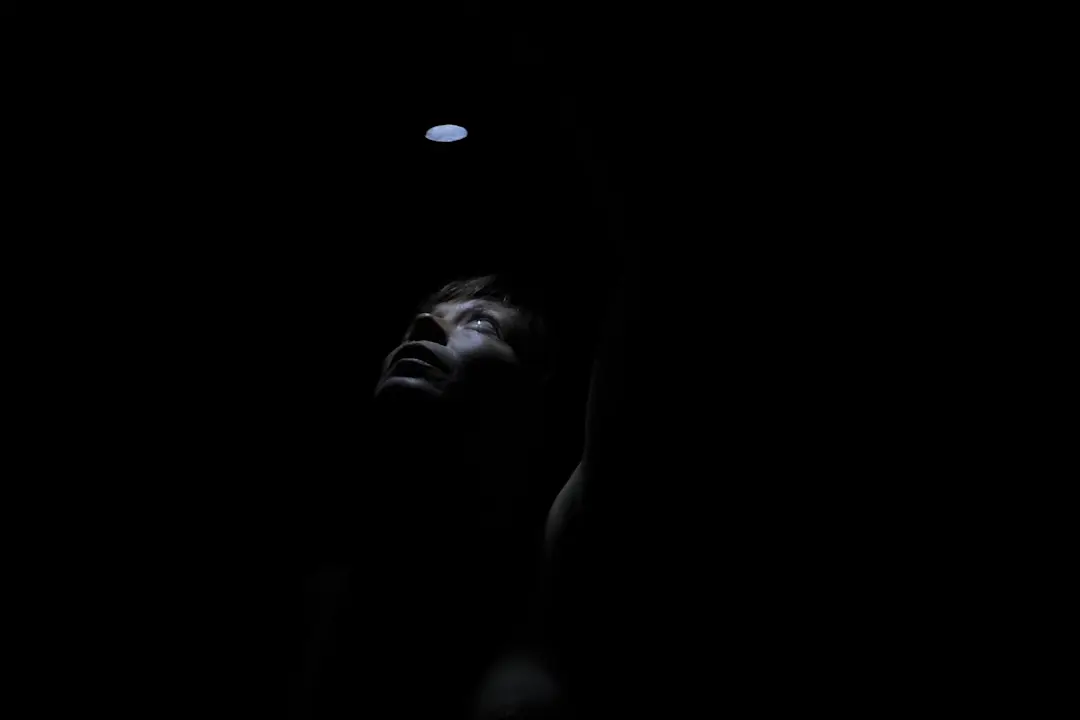 L'Iris en una foscor absoluta, mirant amunt, on una llanterna encesa fa la llum justa per veure-li la cara.