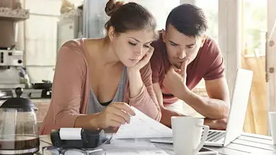 A couple intently reviews documents at a table with a calculator and coffee mugs nearby. A laptop is open, suggesting a focus on financial planning.