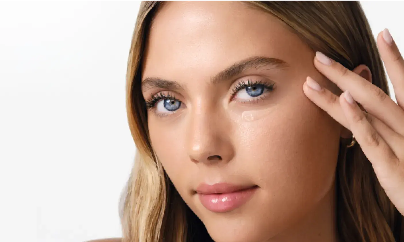 A young woman is applying a clear gel or cream to the skin under her right eye with her fingers.