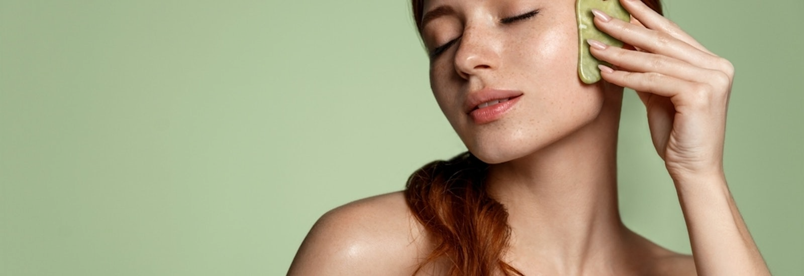 Beautiful redhead woman doing skin care routine after shower, scrapping, massage facial skin with jade gua sha scrapper, body and face beauty concept, green background.