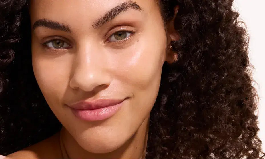 Close-up of a woman with curly hair and natural makeup, smiling softly with glowing skin against a neutral background.