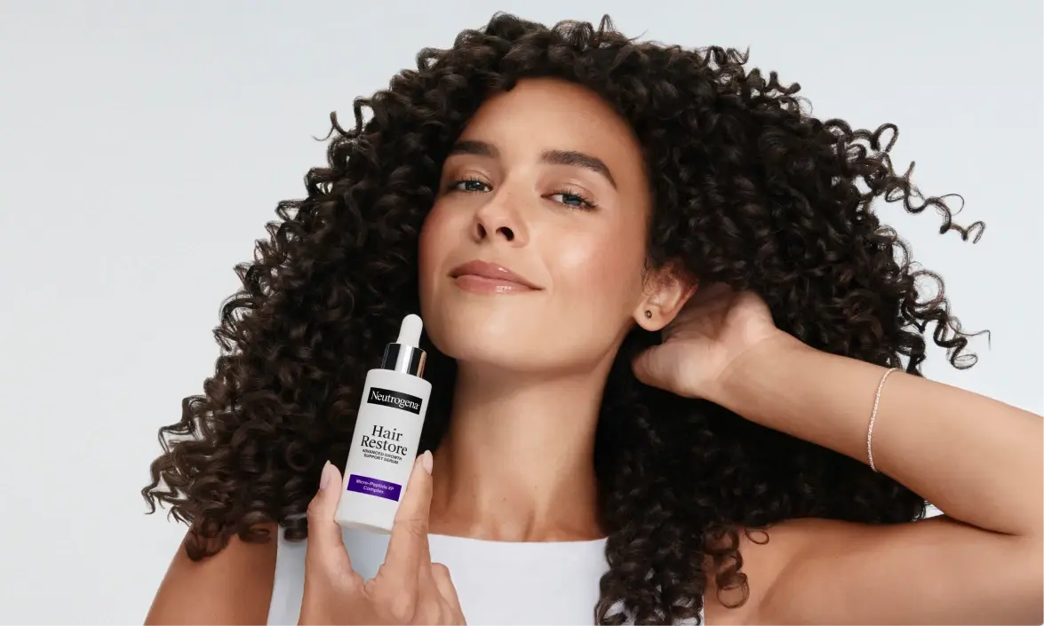 Woman with curly hair holding bottle of Neutrogena® Hair Restore Support Serum