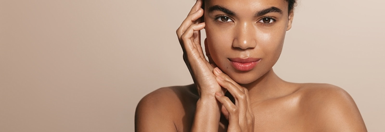 Skin care routine. Dark skinned woman with perfect glowing body, head and shoulders, touches her nourished face after skin cream and moisturizer, brown background