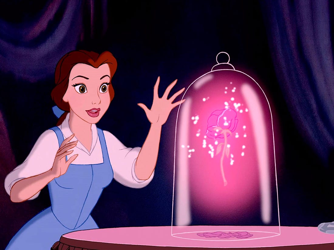 Belle, a white young woman with brown hair and a blue hair bow, wearing a blue dress with a white collared undershirt reaches towards a glass jar placed on a table.  Inside the jar, a glowing pink rose is floating, and several petals have dropped to the table.