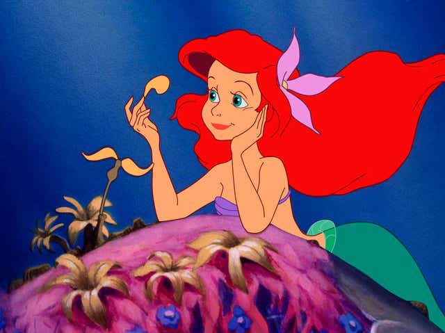 Ariel, a white mermaid with red hair, wearing a purple shell bikini top, with a green fin, picks a yellow flower petal off of a flower growing on a pink coral hill.