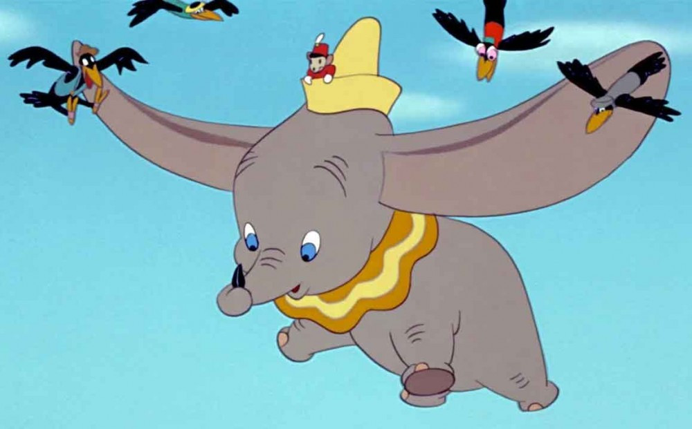 Dumbo, a gray baby elephant with very large ears, wearing an orange and yellow collar and a yellow hat, is flying through the air with a black feather curled in his trunk.  He is surrounded by four Black crows, Fats, Deacon, Dopey, and Specks.