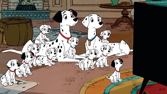 10 black-and-white Dalmatian puppies (all with spots, some with black ears) and two adult Dalmatian dogs (Pongo, with a red collar and black ears, and Perdita, with a blue collar and white ears with spots) sit on the carpet in a living room watching a TV.  There's an orange, yellow, blue and red circular carpet under the television and a brown pouf and brown tea table behind the dogs.