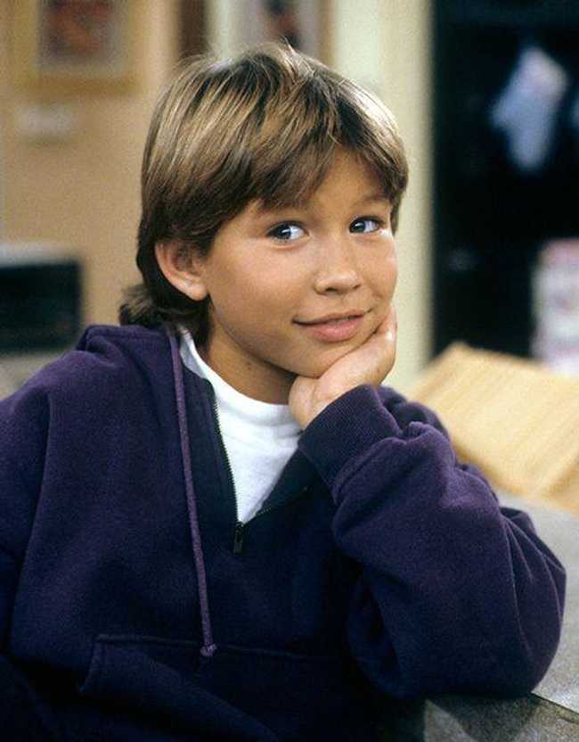 Jonathan Taylor Thomas, a young white actor with blue eyes and blonde hair down to his chin looks off camera, his hand under his chin, wearing a white t-shirt with a dark blue v-neck hooded sweater over it, smiling.