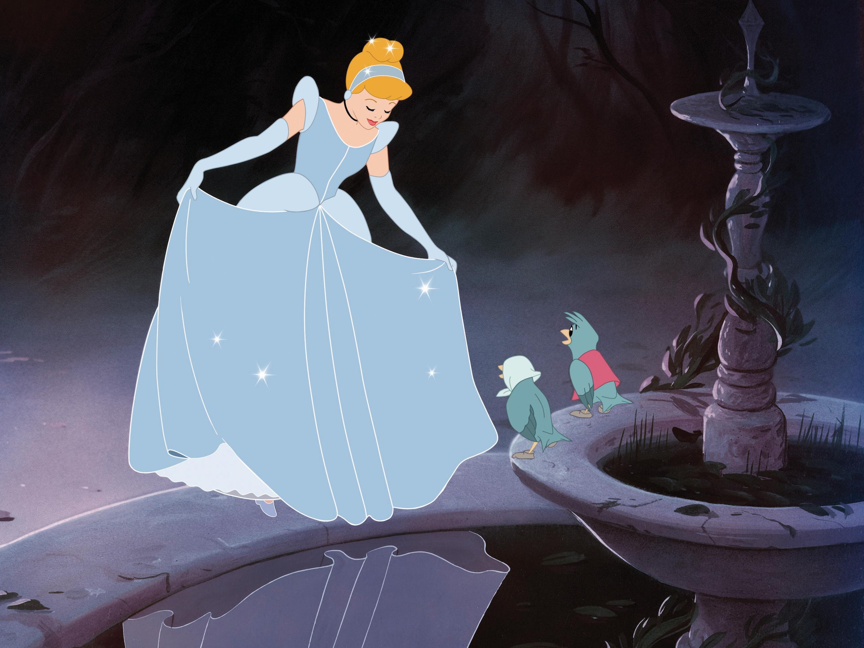 A white woman (Cinderella) in a blue ball gown with sparkles on the skirt stands on the edge of a fountain, looking at her reflection.  Two blue birds (one with a white handkerchief on its head and one with a red vest) watch her.