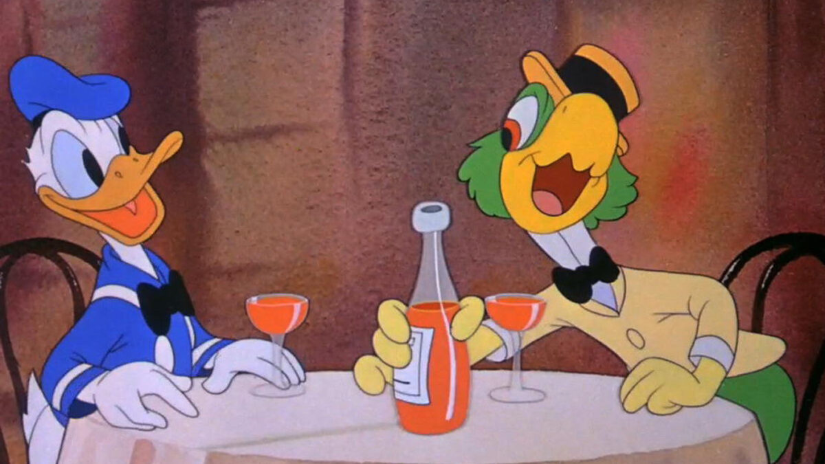 Donald Duck, a white duck with a blue sailor suit and hat on, sits at a table sharing wine with José Carioca, a green parrot wearing a jacket, high collar, bow tie and straw hat with a black band.