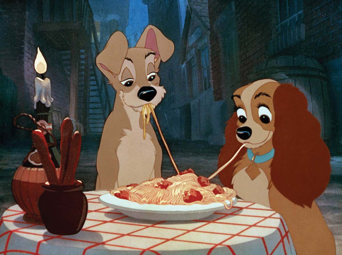 Two dogs, a gray mutt (Tramp) and a brown Cocker Spaniel with a blue collar and gold tag (Lady) sit at a table in an alleyway, eating spaghetti and meatballs.  The table has a lit candle in a wine bottle and a jar of breadsticks.