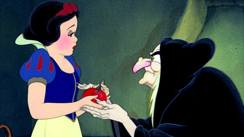 Snow White, a young white girl with black hair, a red hair bow, and a blue, red and yellow dress, holds a red apple in her hand.  An extremely old white woman with a wart on her reddening nose, white hair, and a black cloak, holds Snow White's hands in herss.