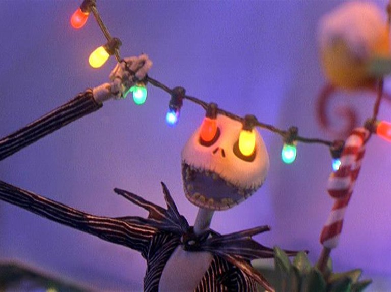 Jack Skellington, a skinny skeleton in a striped suit, holds a string of Christmas lights in front of him, in such a way that two lights, one yellow and one red, are in front of his empty eye sockets.  He is smiling widely, showing many teeth.