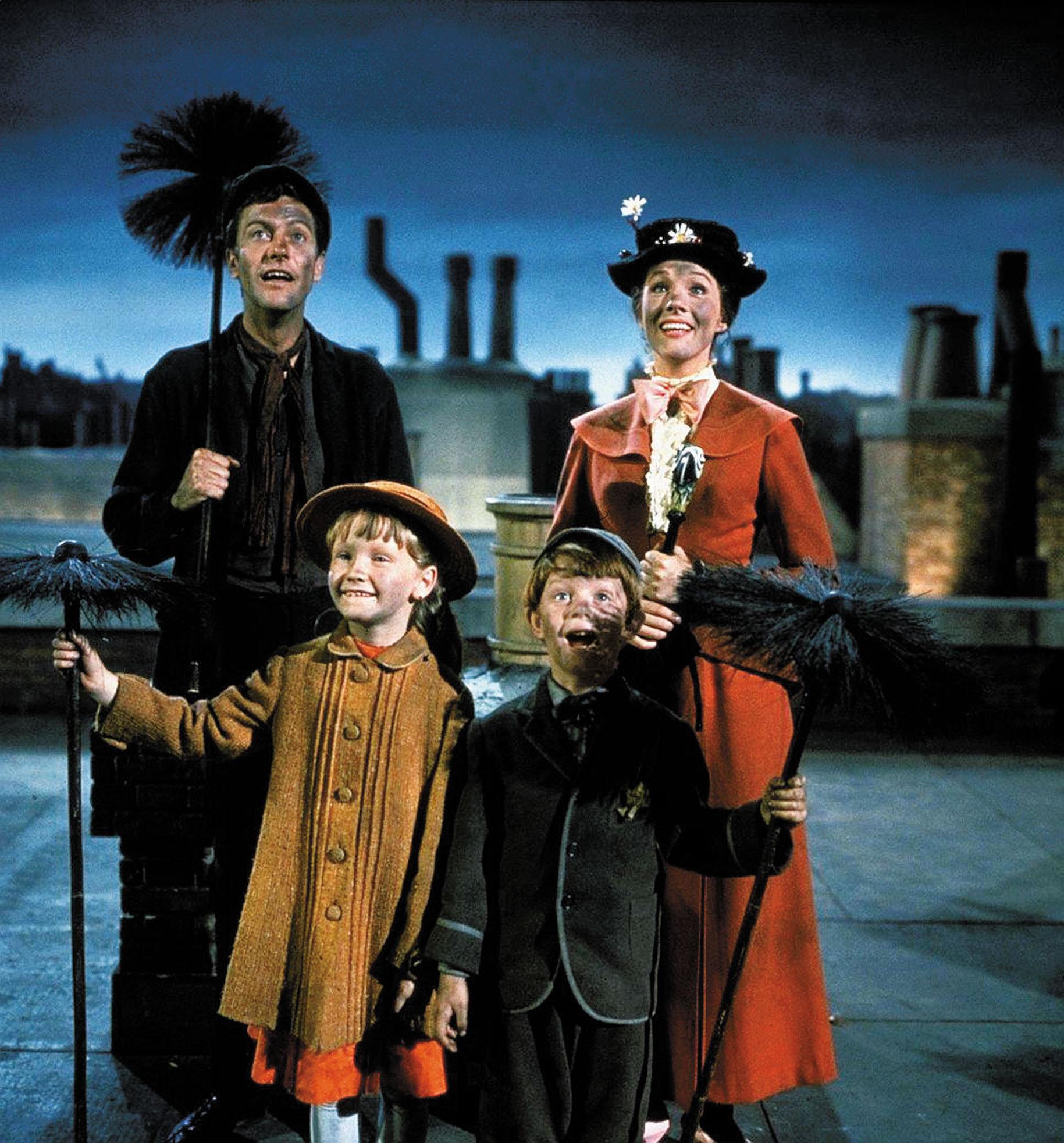 Bert, a chimney sweep, Jane, a young blonde child in a red dress and yellow overcoat, Michael, a young strawberry blonde child in short pants and a button coat, and Mary Poppins, a nanny in a red dress with a black hat with white flowers, stand on a rooftop, covered in soot.