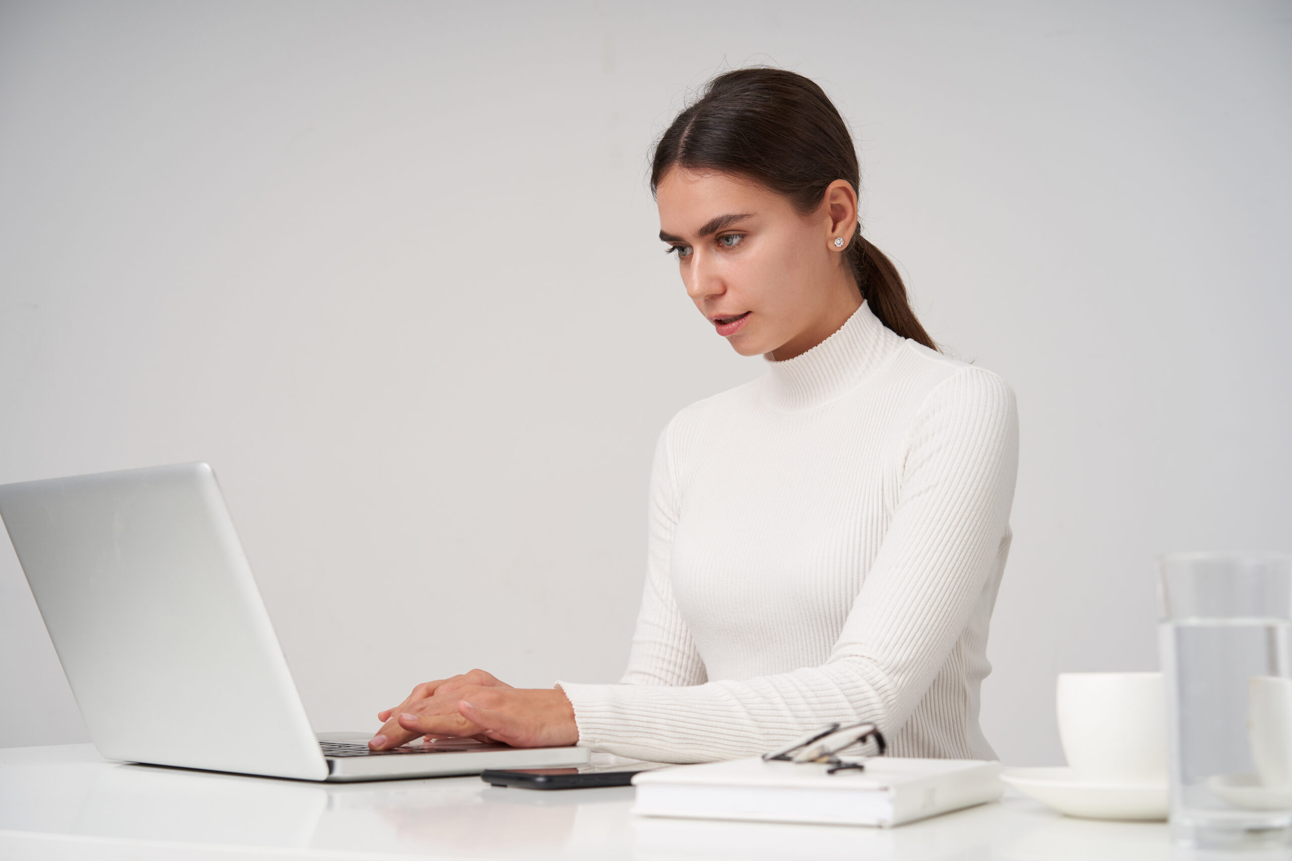 portrait-young-dark-haired-businesswoman-white-knitted-poloneck-sitting-white-wall-with-laptop-keeping-hands-keyboard-looking-screen-seriously-scaled