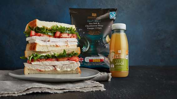 Premium meal deal asset - roast chicken sandwich stacked on plate, sea salt & chardonnay wine vinegar crisps and cloudy apple juice on table with dark background. 