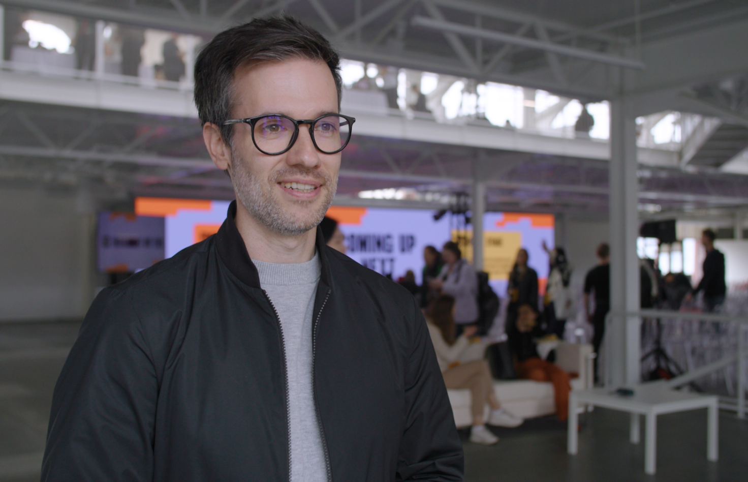 Person with glasses and dark jacket smiling at a conference venue with colorful displays in the background.