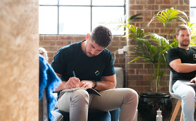 Person in dark shirt writing in notebook in modern brick office space with plants and large windows.