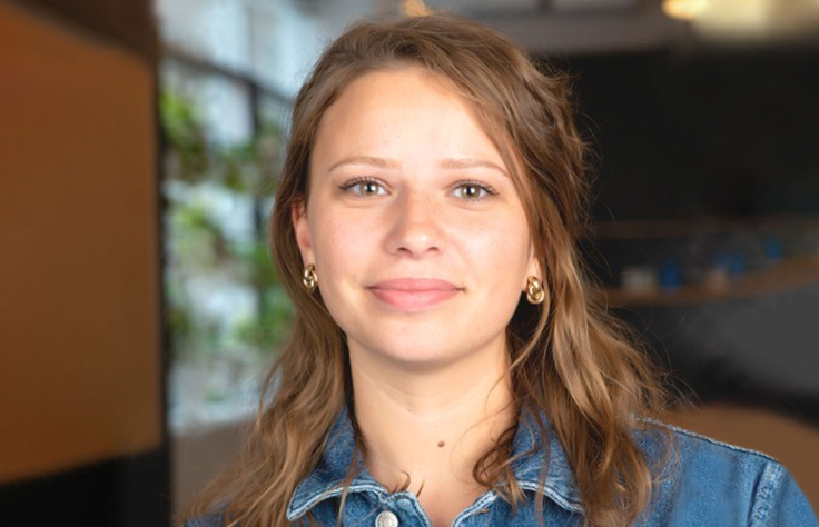 Person with brown hair wearing denim jacket in indoor setting with blurred background and natural lighting.