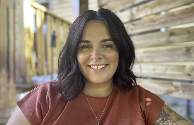 Person with dark shoulder-length hair smiling at camera, wearing rust-colored top against wooden wall background.