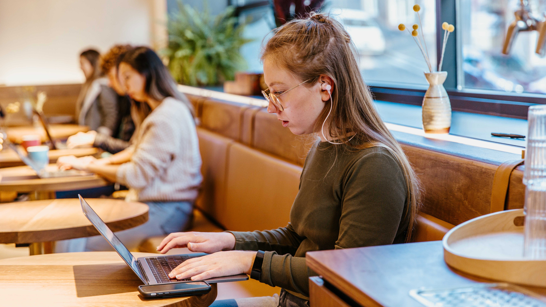 Person with earbuds working on laptop in cozy café with brown leather seating and natural light.